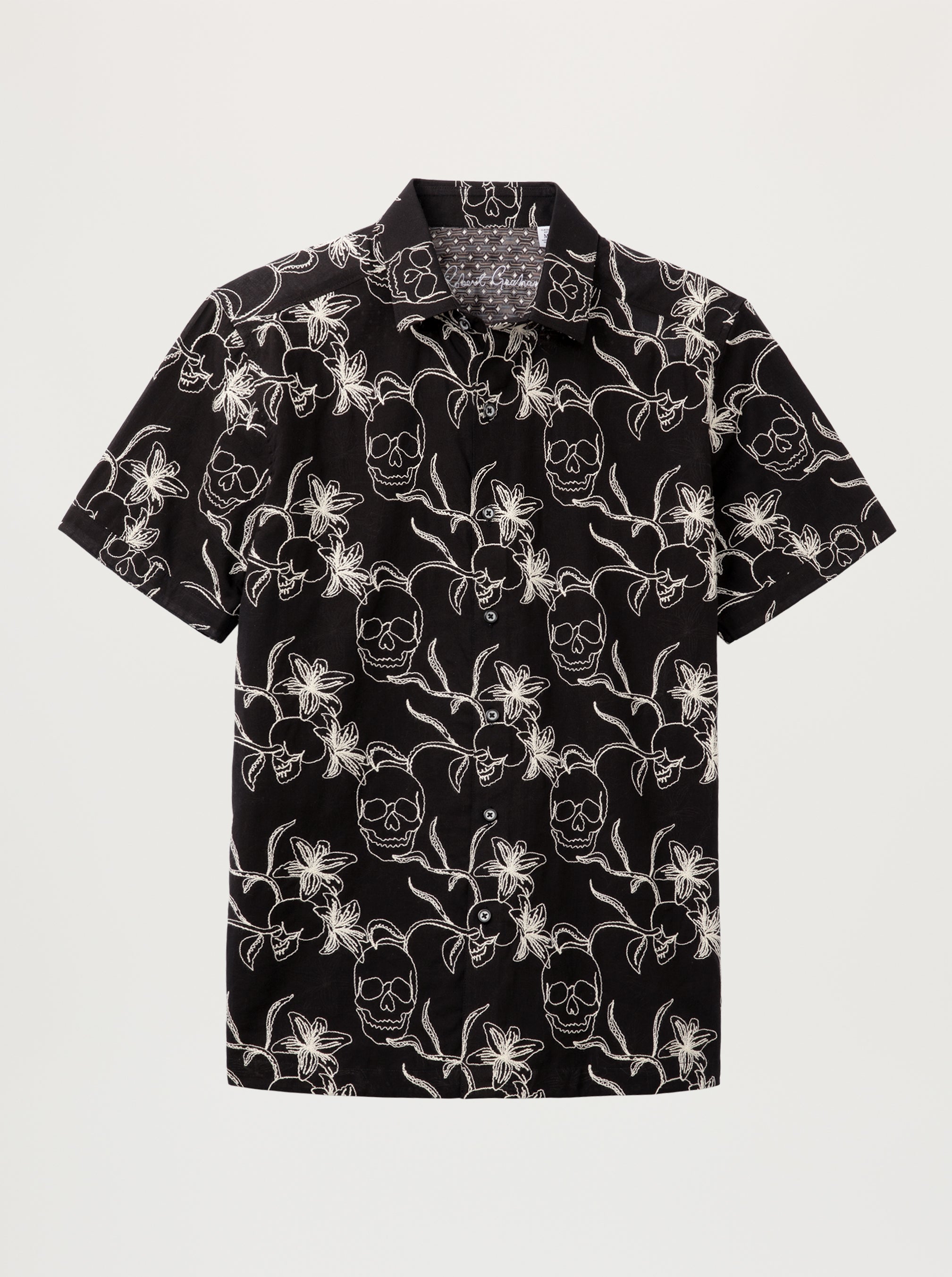 A short-sleeve button-up shirt with a black base and an all-over white skull and floral pattern, displayed flat against a light gray background.
