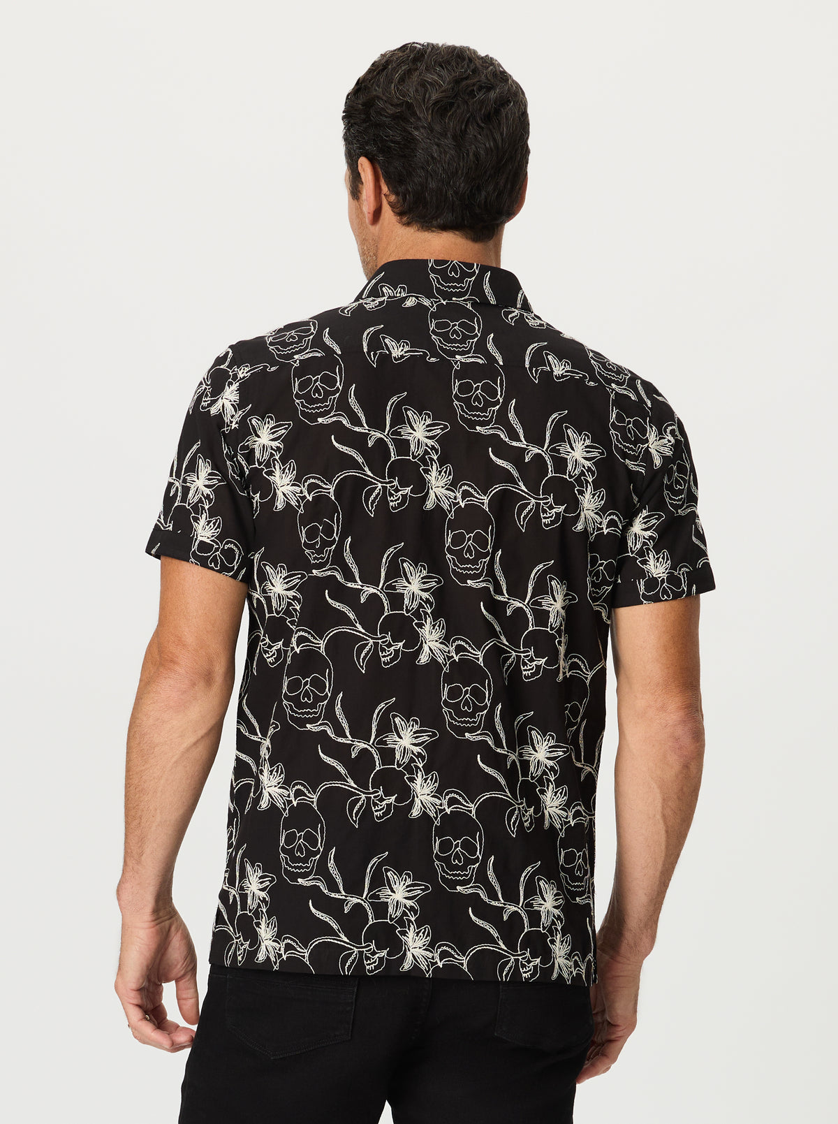 Man wearing a black shirt with white florals and skulls pattern on a plain background