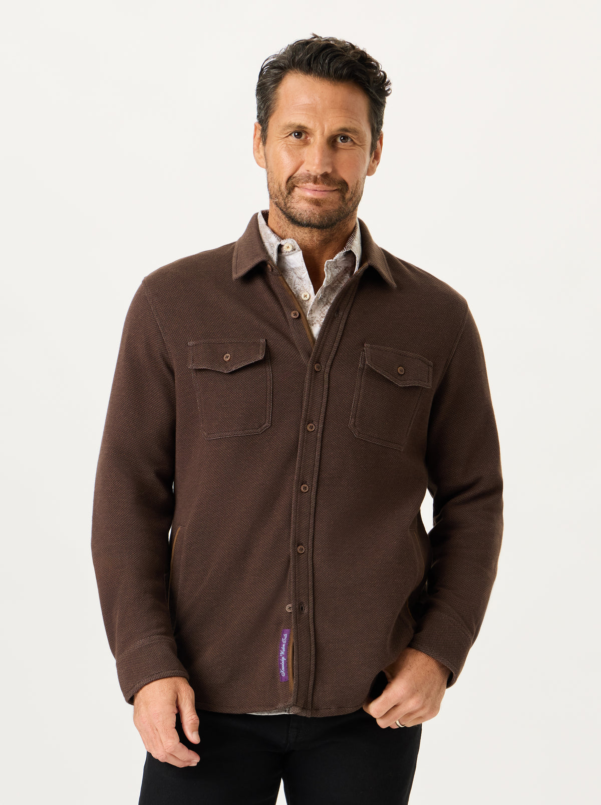 A man wearing a dark brown long-sleeve button-up shirt jacket over a patterned collared shirt, paired with black pants, photographed against a clean white studio background.