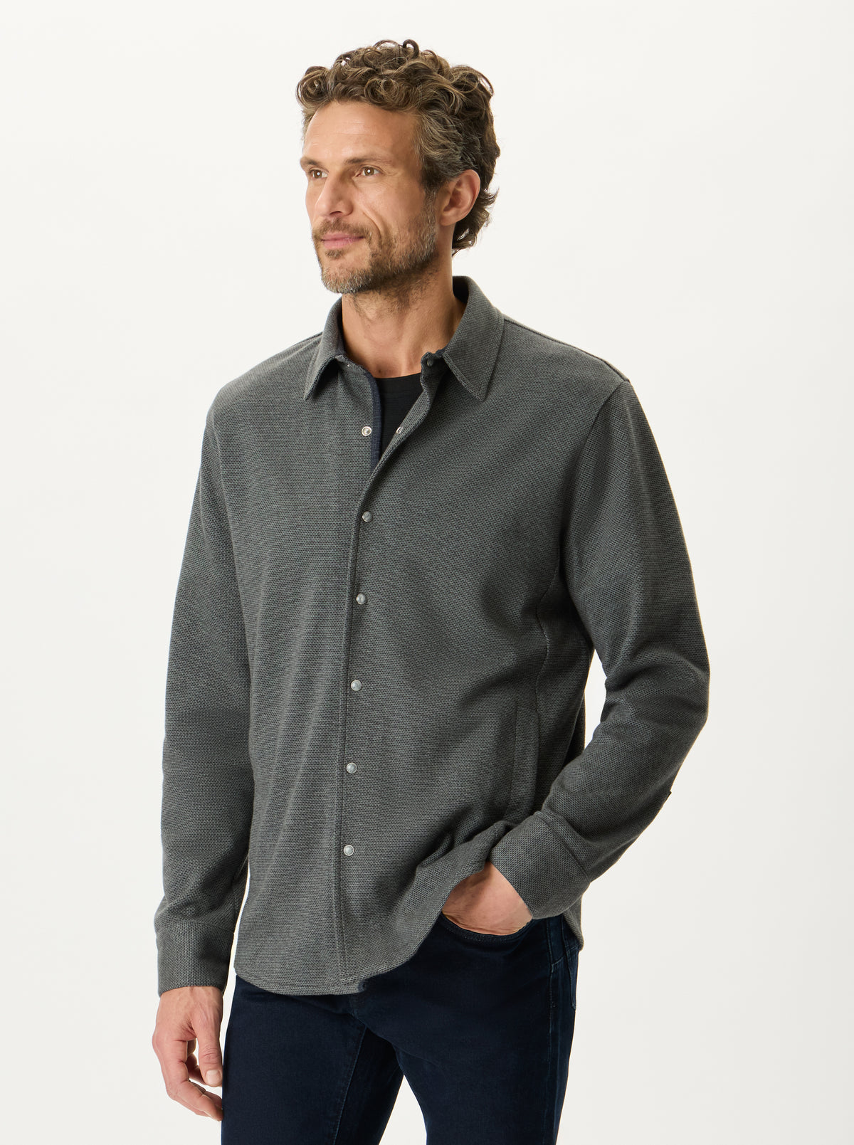 A man wearing a dark gray long-sleeve button-up shirt over a black undershirt, paired with dark blue jeans, photographed against a clean white studio background.