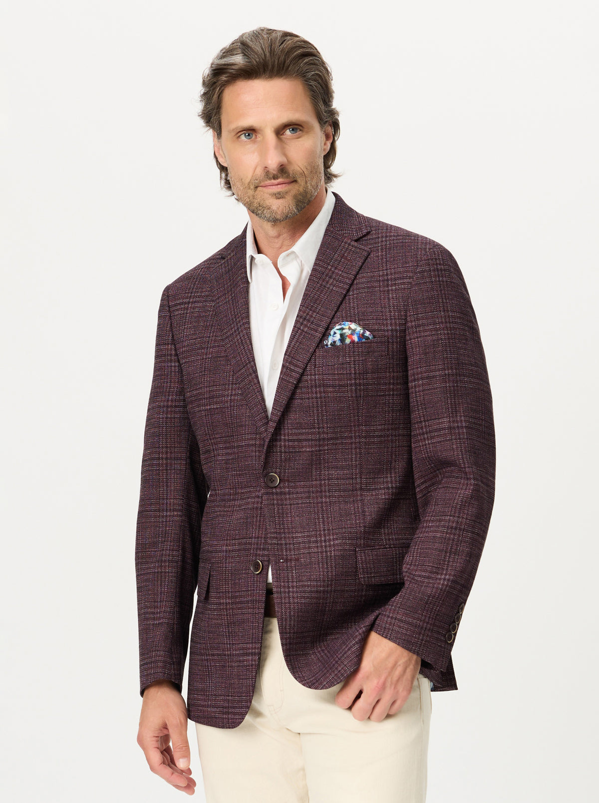 A man wearing a dark plum-colored plaid blazer over a white dress shirt and cream trousers, standing against a plain white studio background.