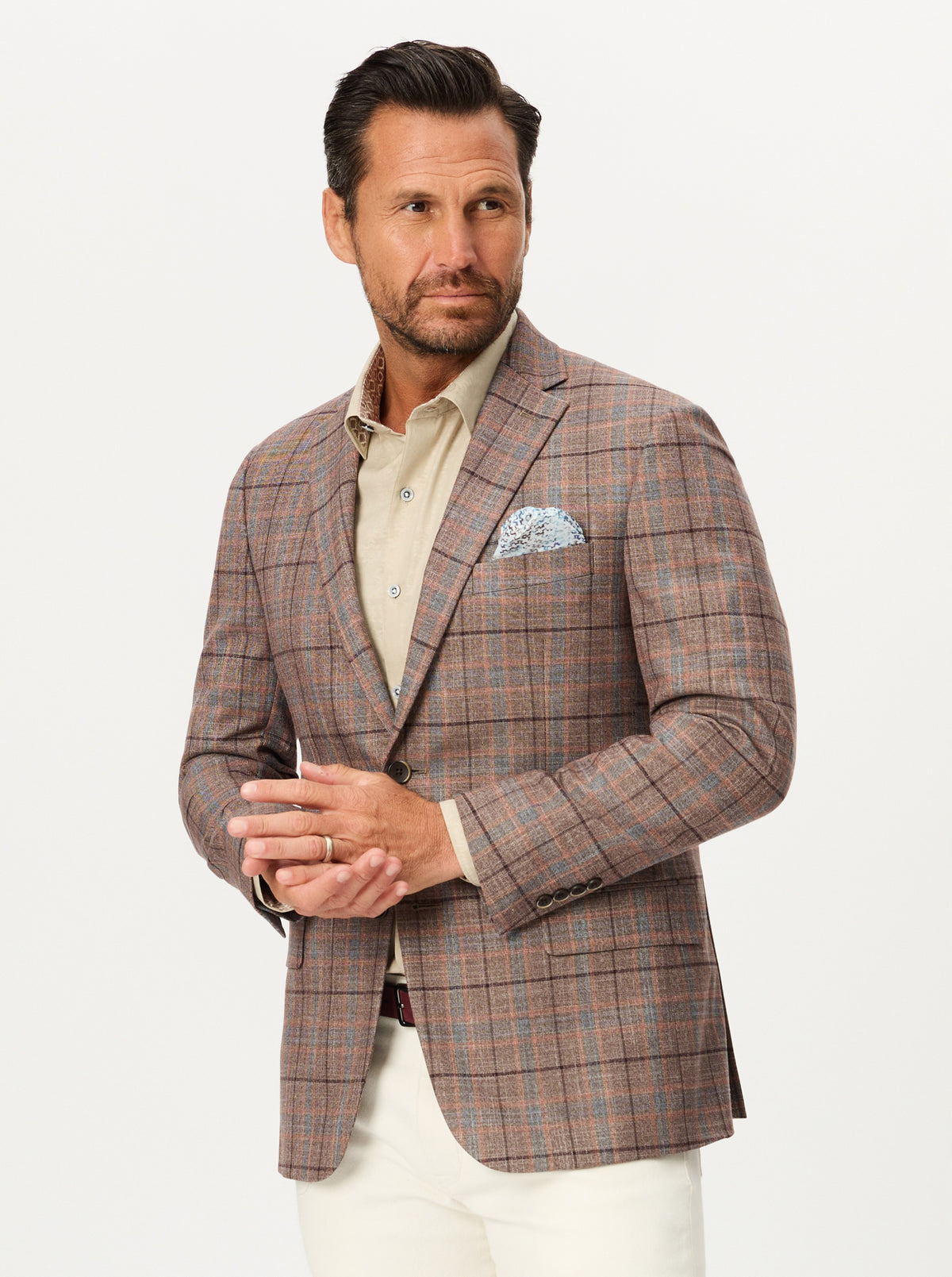 A man wearing a brown and beige plaid blazer over a light beige shirt, styled with white trousers and a patterned pocket square, photographed against a plain light gray background.