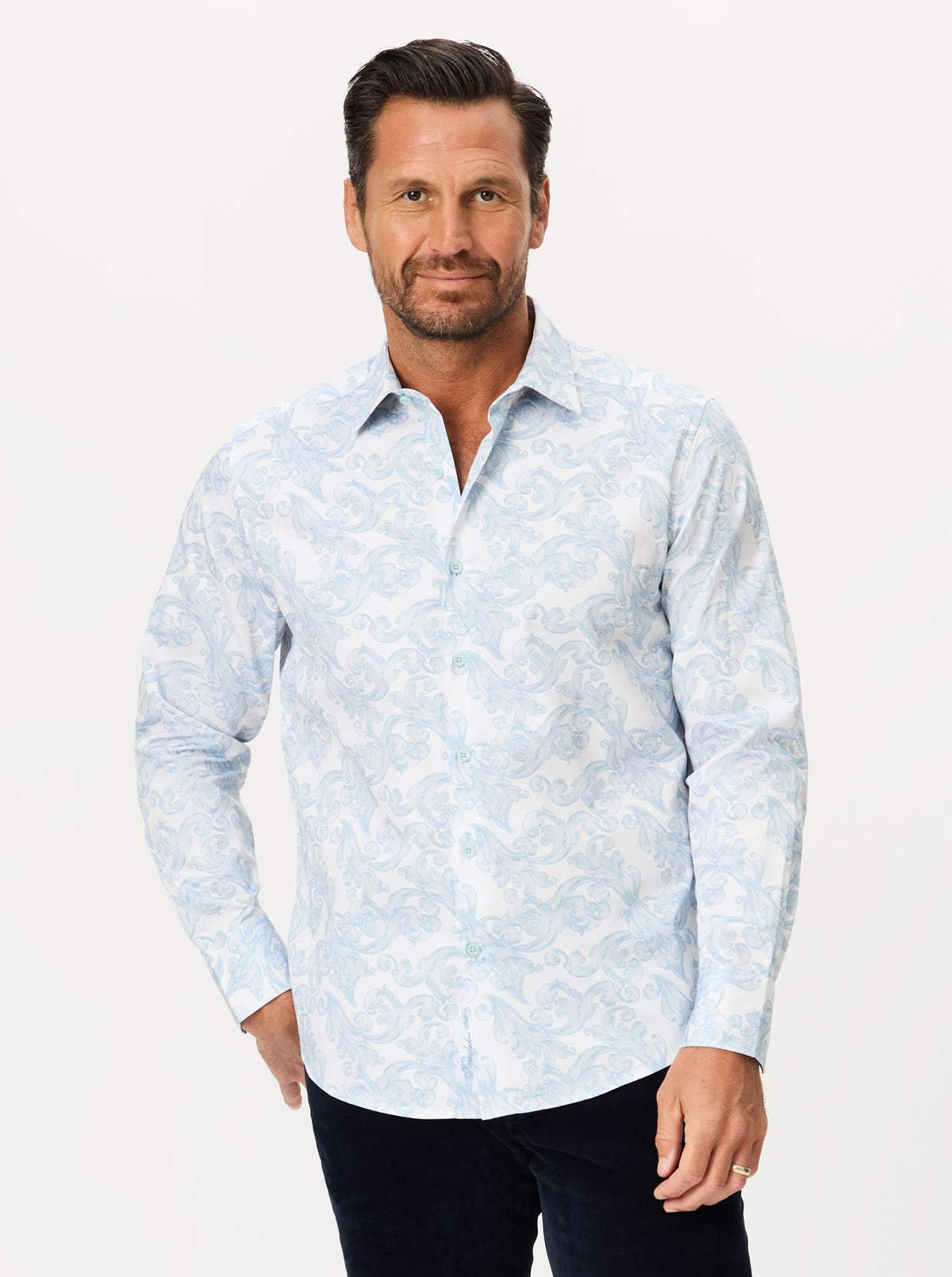 A man wearing a light blue and white patterned long-sleeve button-down shirt, paired with dark pants, standing against a plain light background.