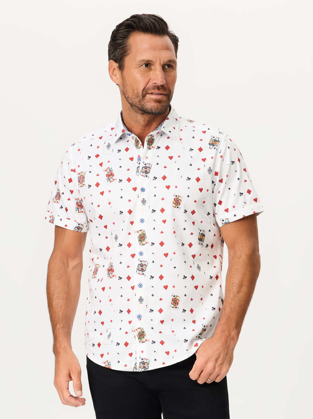 A man wearing a white short-sleeve button-up shirt with an all-over playing card pattern, paired with black pants, against a plain light gray studio background.
