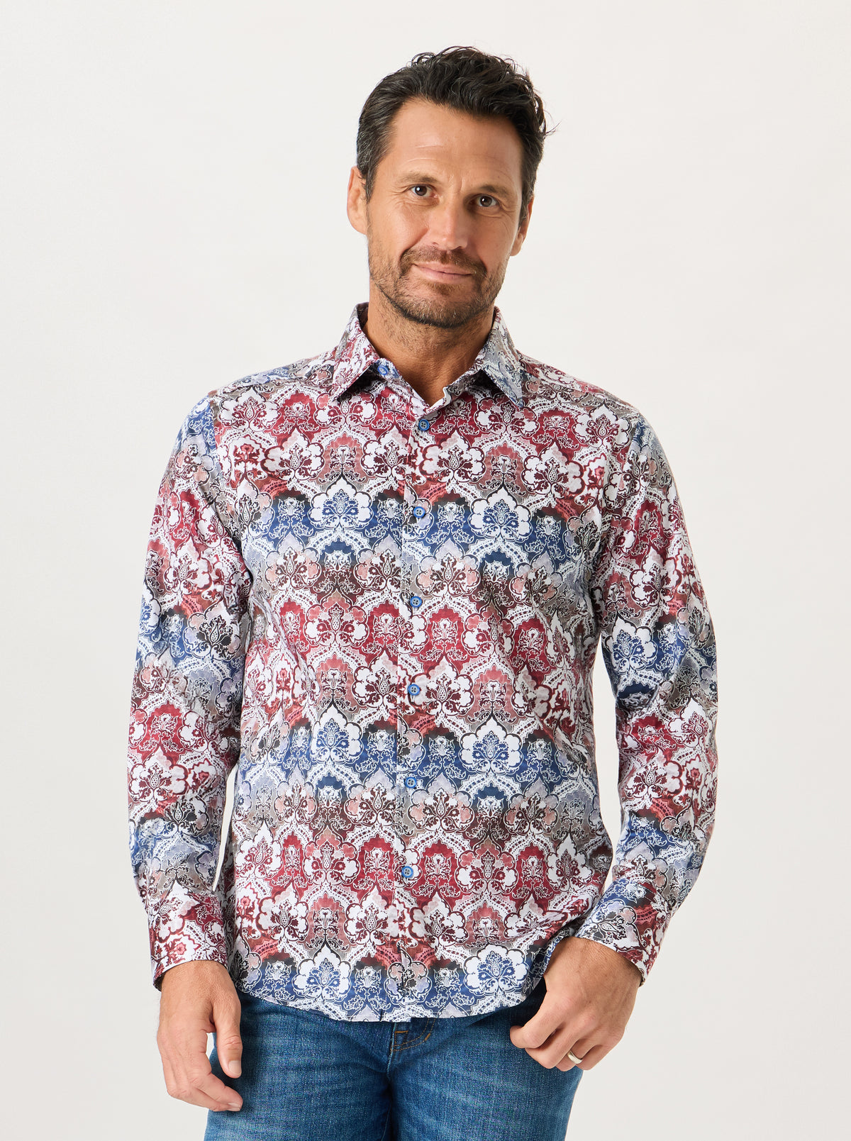 A man wearing a long-sleeve button-up shirt with an intricate red, blue, and white geometric pattern, paired with blue jeans, against a plain light gray background.