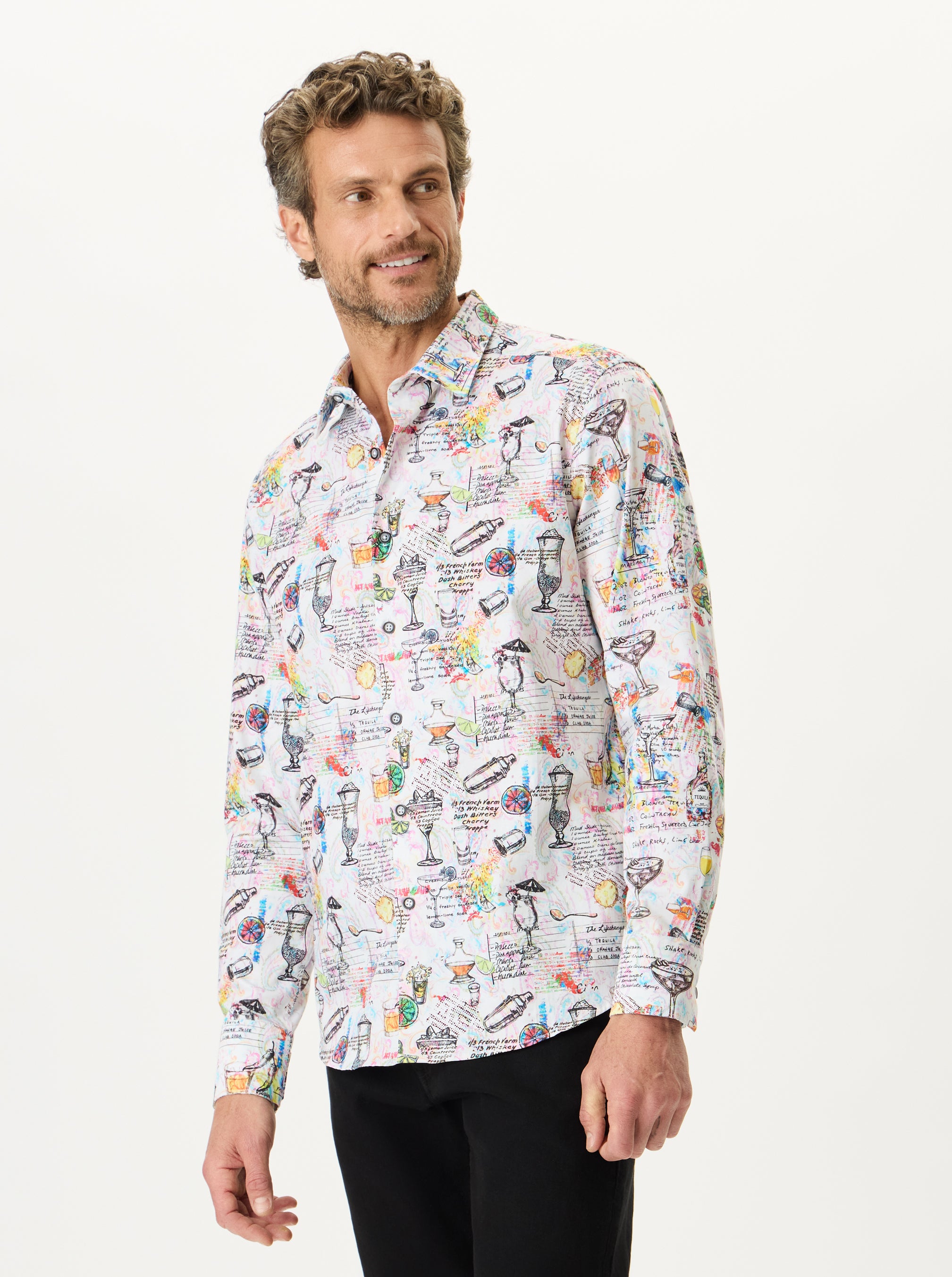 A man wearing a long-sleeve button-up shirt with a dense multicolor graphic print of cocktails, glasses, and handwritten notes on a white base, paired with black trousers, photographed against a solid off-white background.