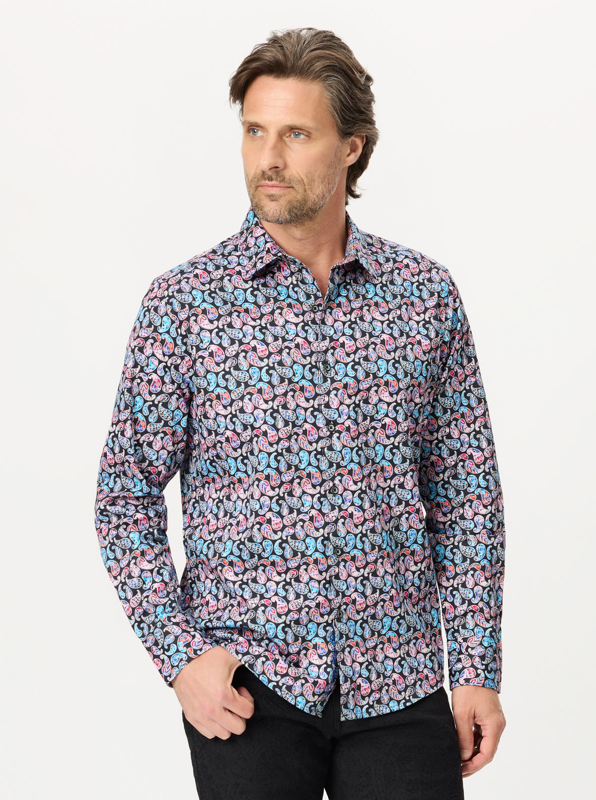 A man wearing a long-sleeve button-up shirt with an all-over abstract pattern in pink, blue, and white, paired with black pants, against a light gray studio background.