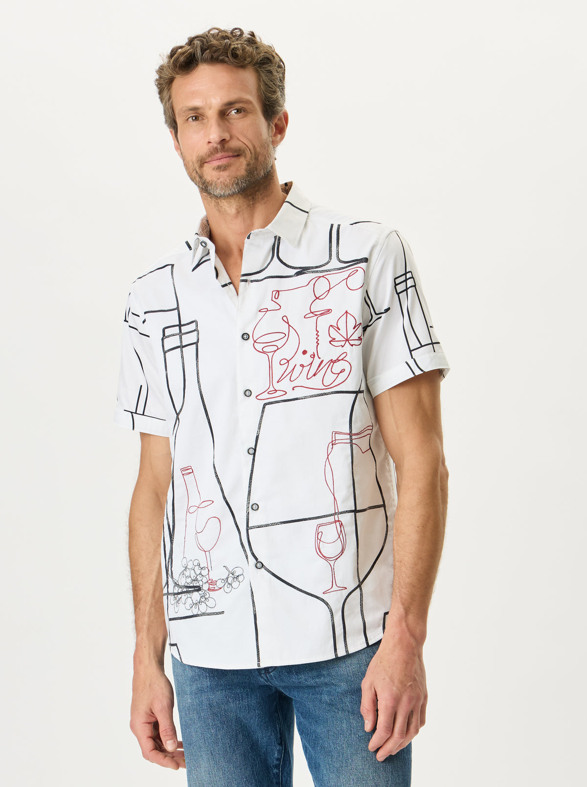 A man wearing a white short-sleeve button-up shirt with a black line-art graphic print of wine glasses, bottles, and the word 'Living' in red script, paired with blue jeans, against a plain light background.