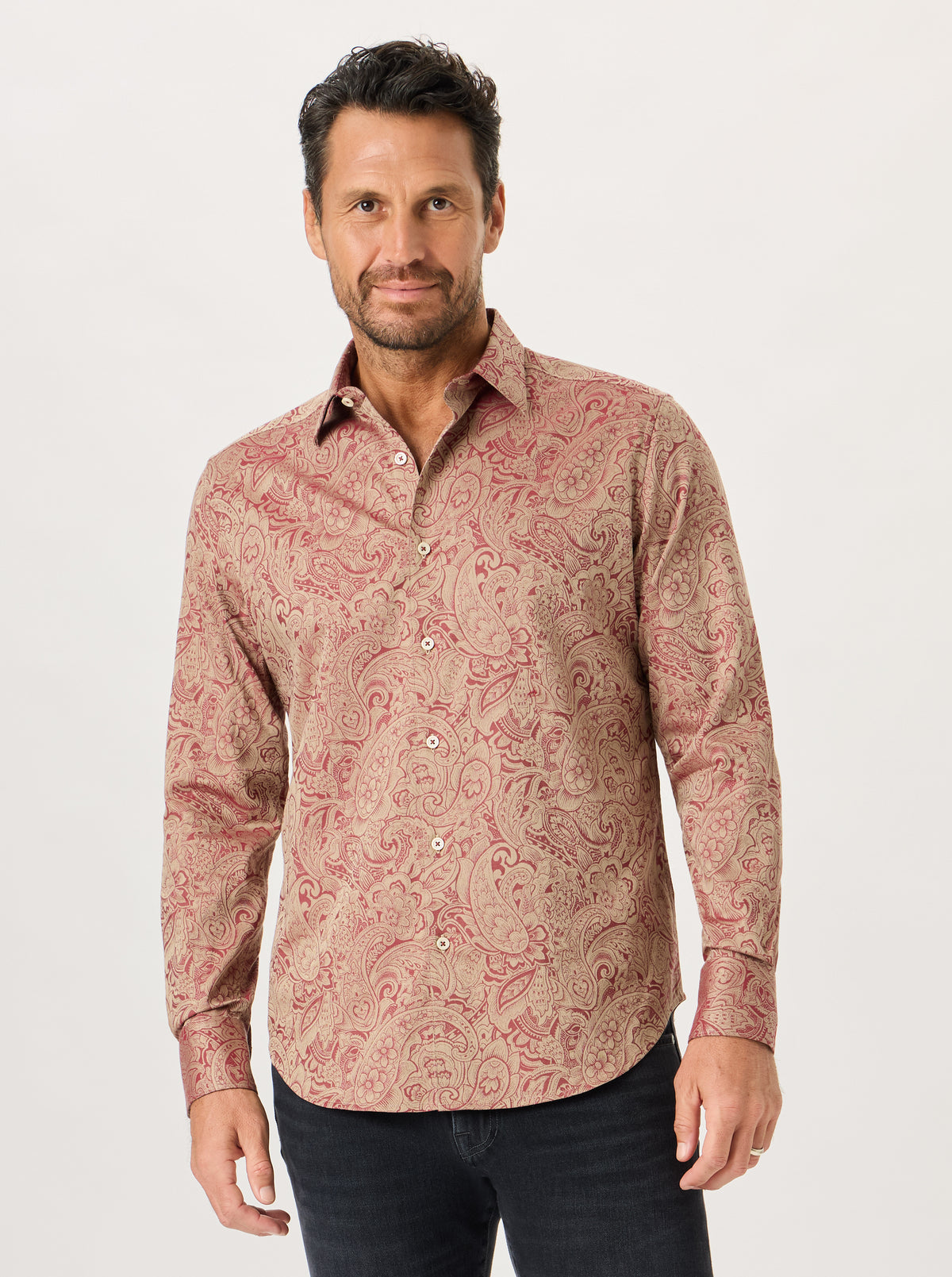 A man wearing a long-sleeve button-up shirt with a beige and dusty red paisley pattern, paired with dark jeans, against a plain light gray background.