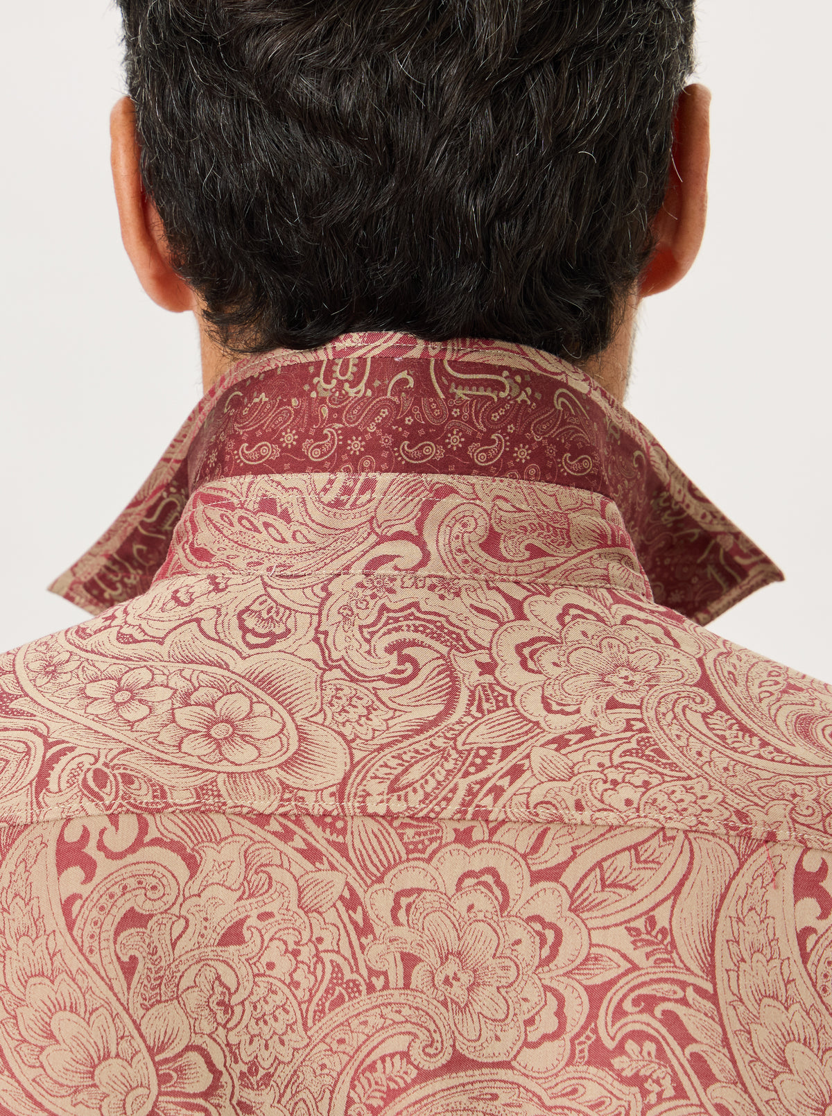 Close-up back view of a man wearing a collared button-up shirt with an ornate paisley and floral pattern in dusty rose and beige.