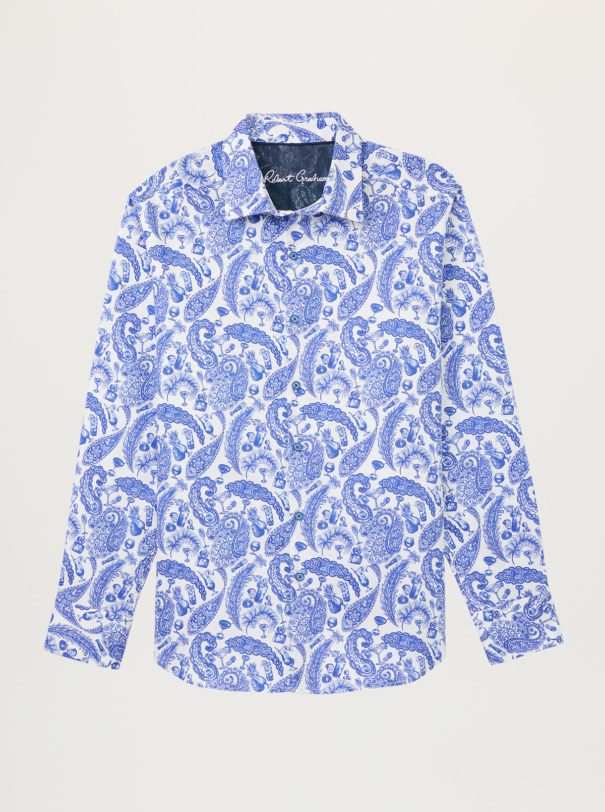 Image of A cotton satin shirt with a paisley print drawn with cocktail glasses, bottles, and pineapples worked into the motifs lying flat on a grey background. 