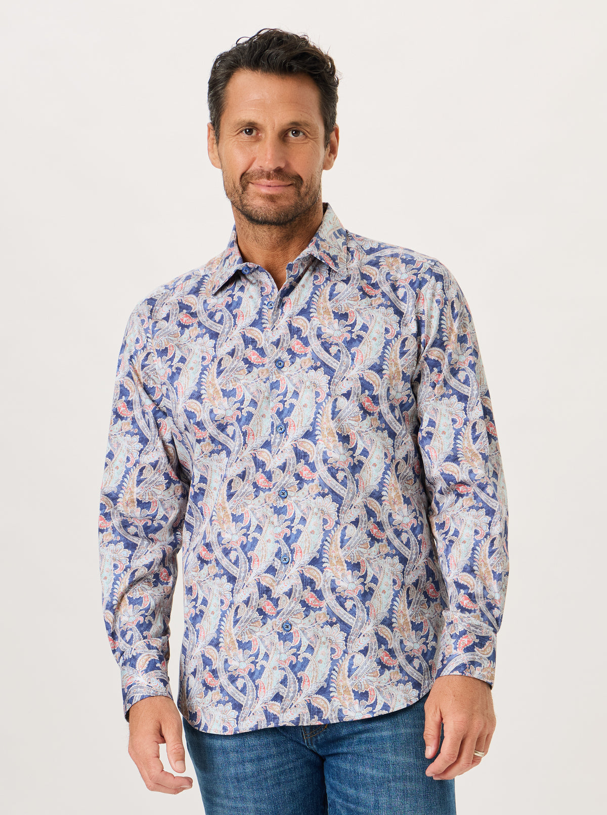 A man wearing a long-sleeve button-down shirt with a blue, white, and pink paisley pattern, paired with blue jeans, photographed against a plain light background.
